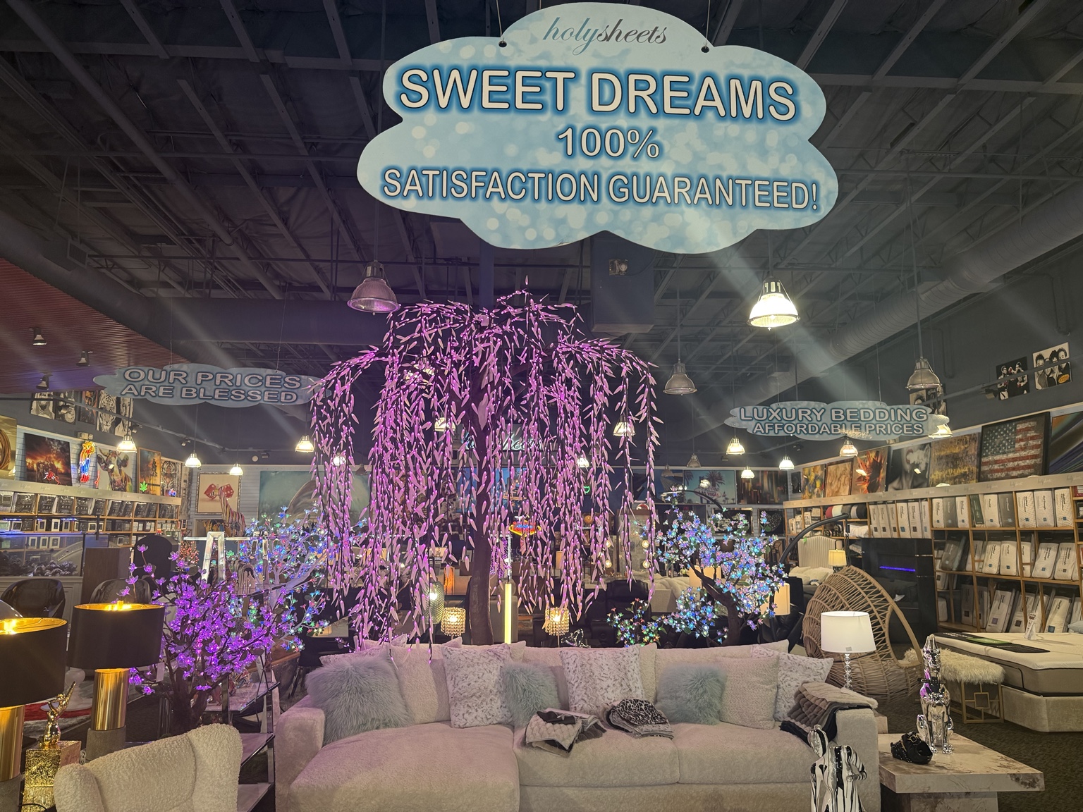 Wide showroom view with white sectional, pillows, beds, wall art, and purple illuminated trees inside HolySheets Galleria