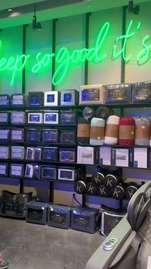 Owner-supplied showroom video screenshot showing packaged bedding and rolled pillows arranged on black shelving beneath green illuminated lettering
