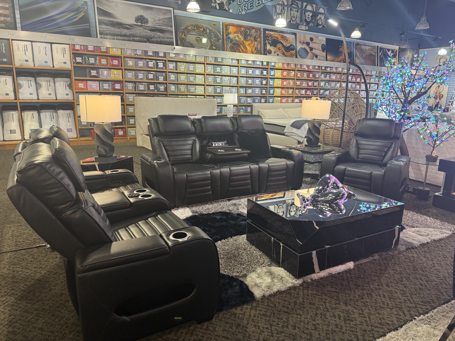 Black massage chairs arranged in front of beds, packaged bedding, artwork, and a lighted decorative tree inside HolySheets Galleria