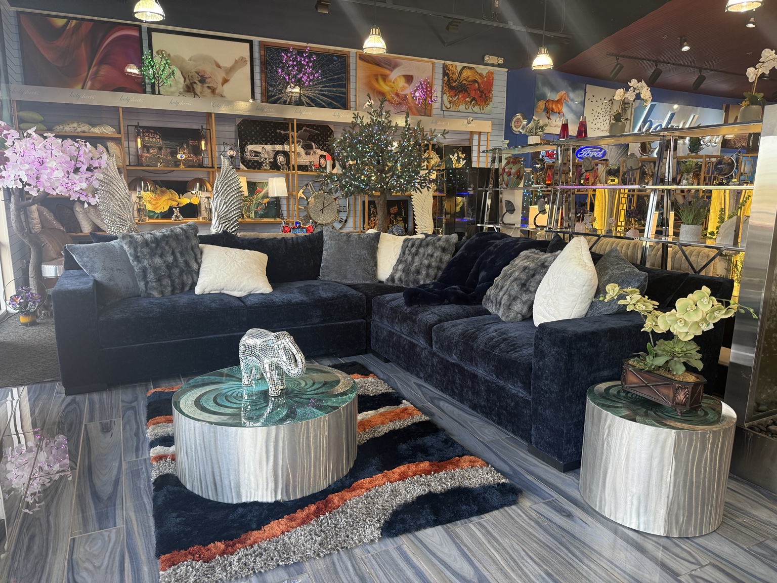 Dark sectional with textured pillows, round accent tables, wall art, illuminated trees, and collectibles inside HolySheets Galleria